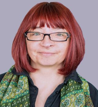 Claudia Hashmi. Eine Frau mit Brille und schulterlangen rotgefärbte Haare. Sie trägt ein Halstuch