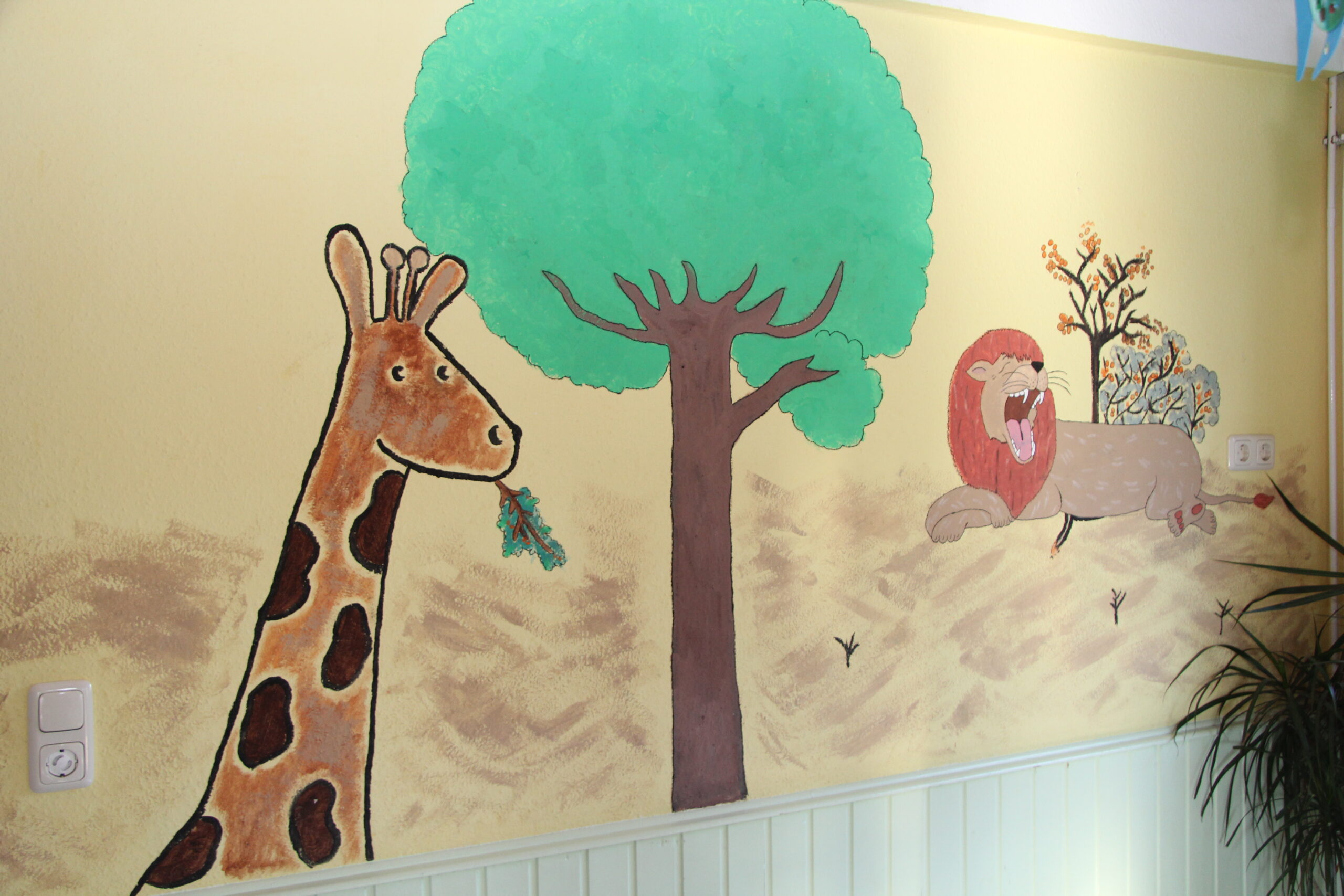 Man sieht im Raum eine gemalte Wand im Raum mit einer Giraffe, einem Löwen und einem Baum