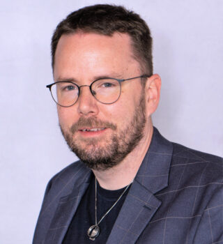 Stefan Flohr. Ein Mann mit Bart, mit einer Brille und kurzen dunklen Haaren. Er trägt eine Kette