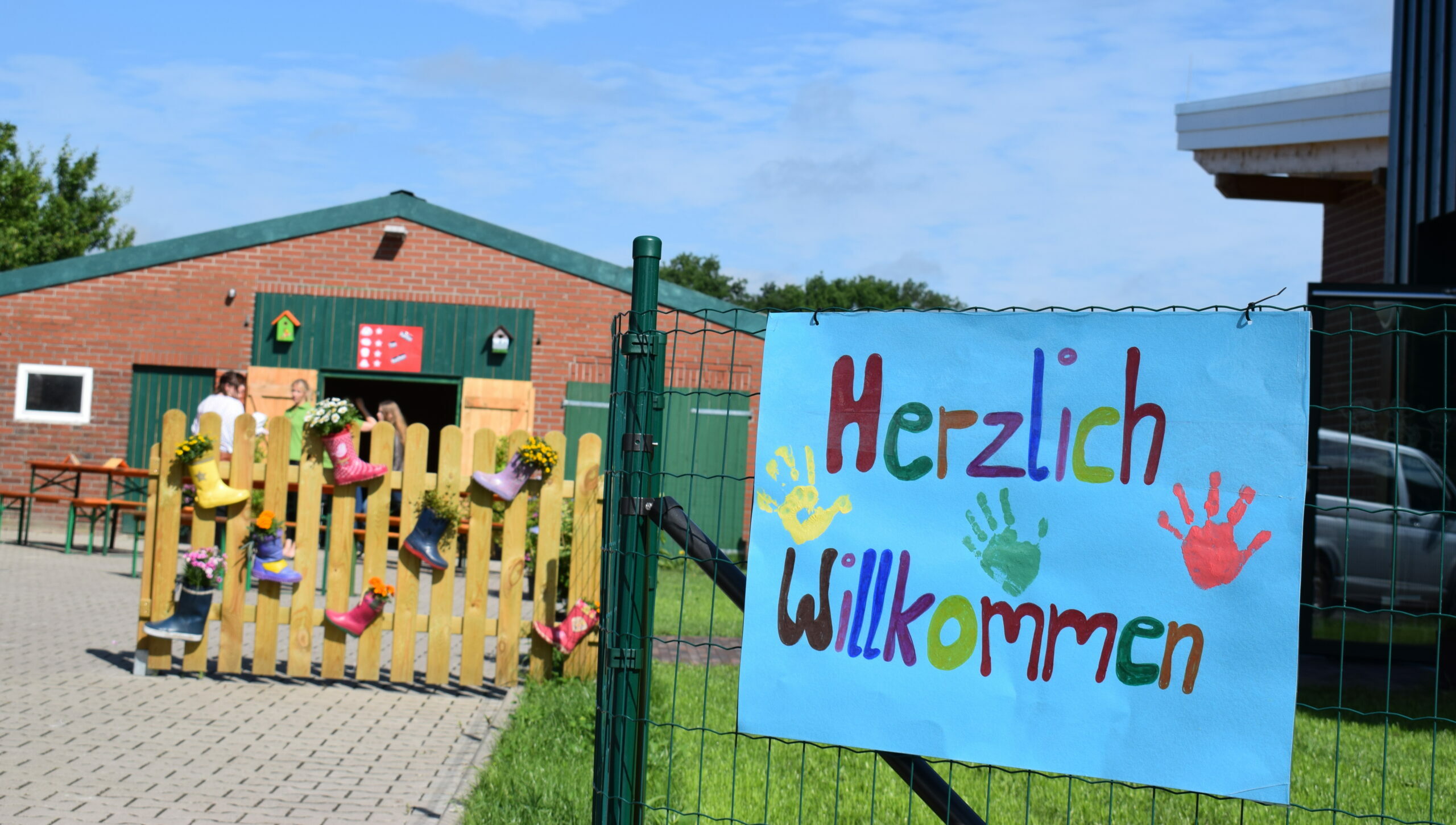 Ein Zaun draußen an der Einfahrt des Kindergartens mit einem Plakat mit der Aufschrift "Herzlich Willkommen" und das Gelände des Kindergartens