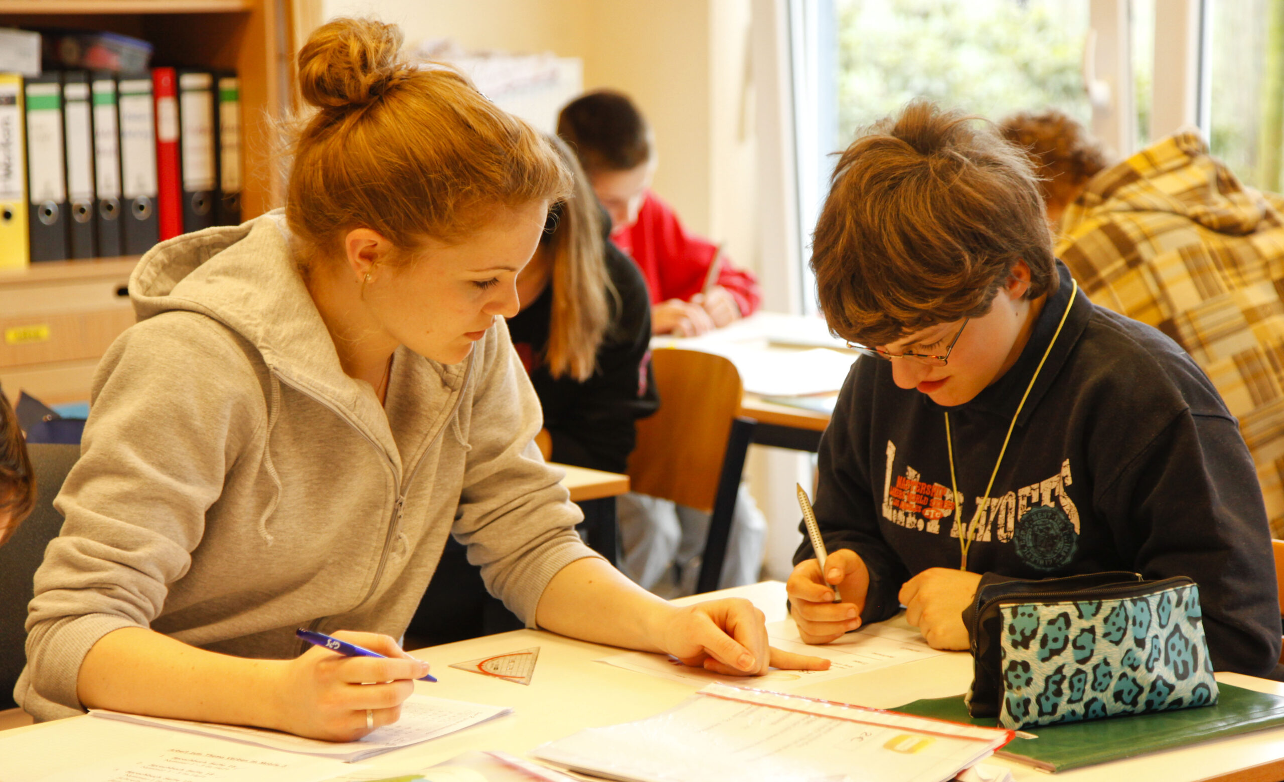 Man sieht im Klassenzimmer Schüler, die ihre Schulaufgaben am Tisch machen. Man sieht auch eine junge FSJ'lerin, die einem Schüler bei seiner Schuaufgabe hilft