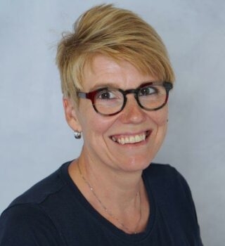 Nicole Ehmen. Eine lächelnde Frau mit kurzen blonden Haaren und einer Brille. Sie trägt Ohrringe