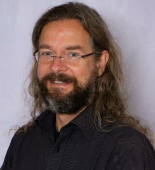 Kay Engel. Ein lächelnder Mann mit einer Brille. Er hat lange dunkle Haare und einen Bart