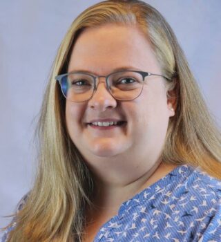 Nadine Dirks. Eine lächelnde Frau langen blonden Haaren und mit einer Brille. Sie trägt Ohrringe