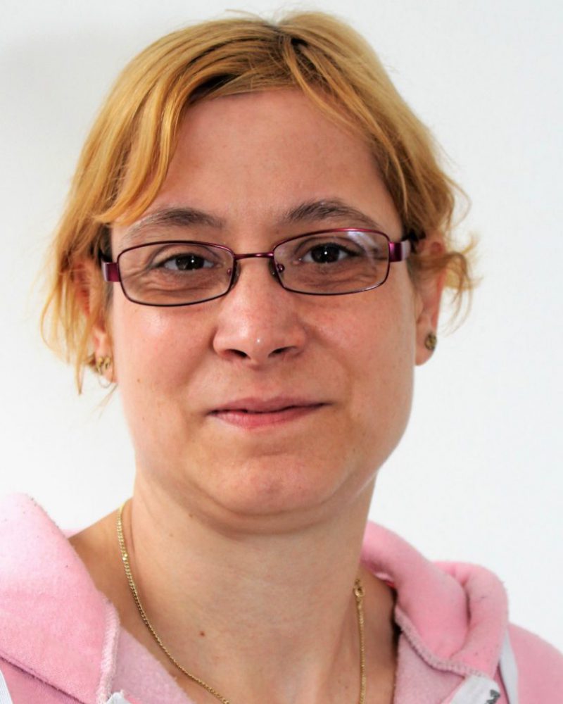Eine Frau mit kurzen rötlichen Haaren. Sie trägt eine Brille und eine Kette