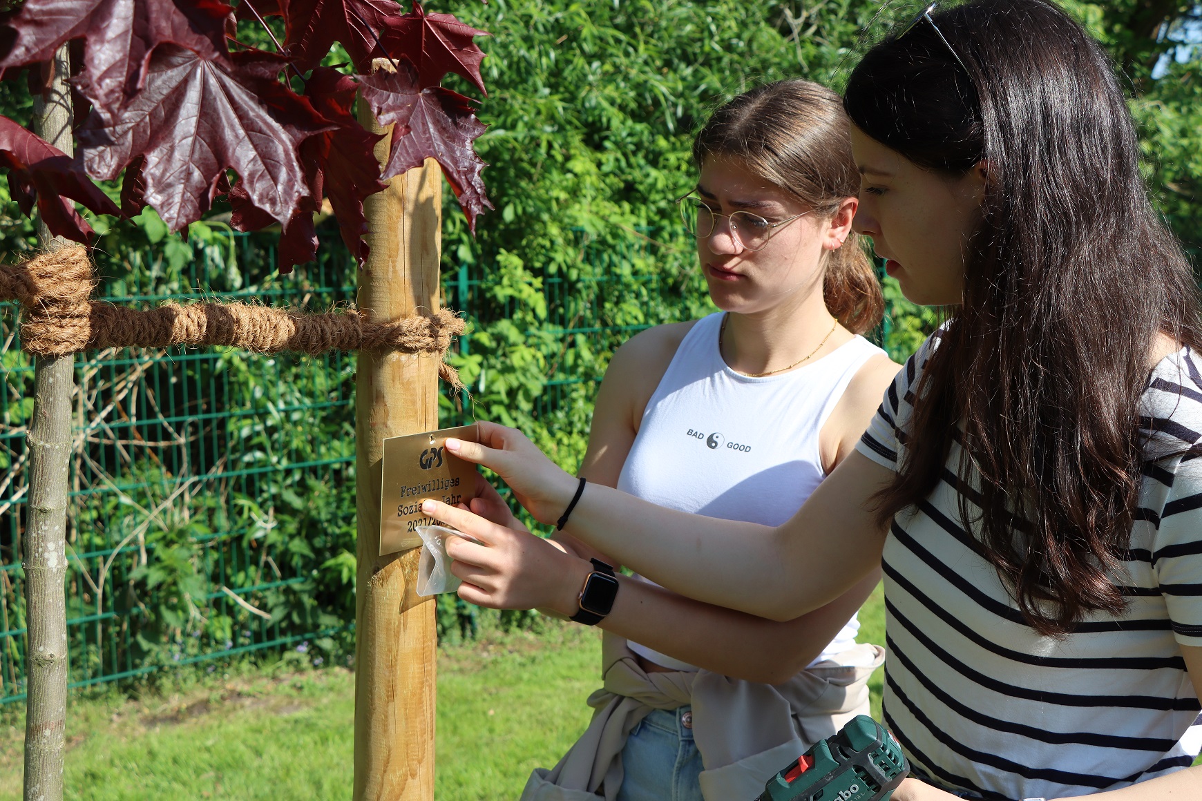 Zwei FSJlerinnen halten ein "GPS Freiwillig Soziales Jahr 2021/2022" an den Mast des Baumes